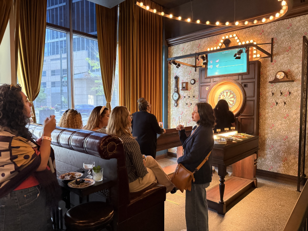 A group of people socializing in a stylish bar with a dartboard, drinks, and warm lighting.