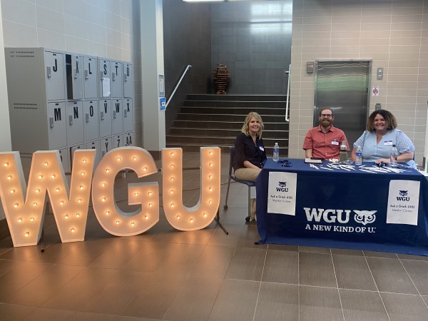 A WGU information booth featuring large light-up letters and three staff members engaging with visitors.