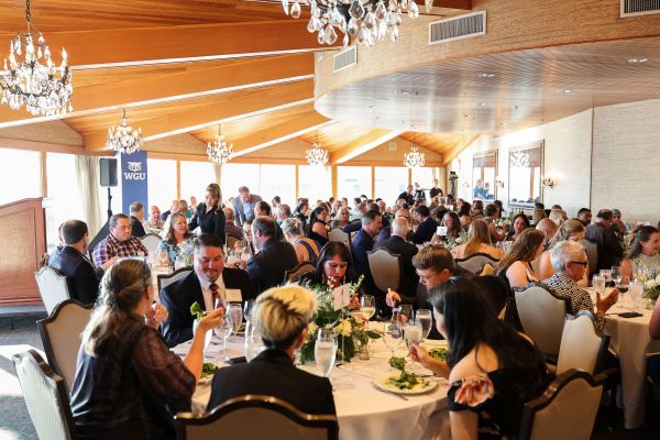 A large group of people attending a formal dinner or event in a spacious room with elegant chandeliers and wooden beams.