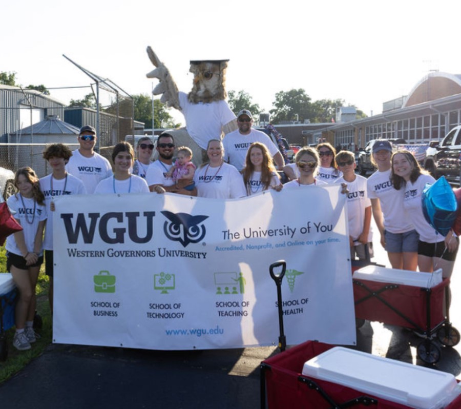A group of diverse individuals, including adults and children, and a large owl mascot, stand behind a prominent white banner for Western Governors University.