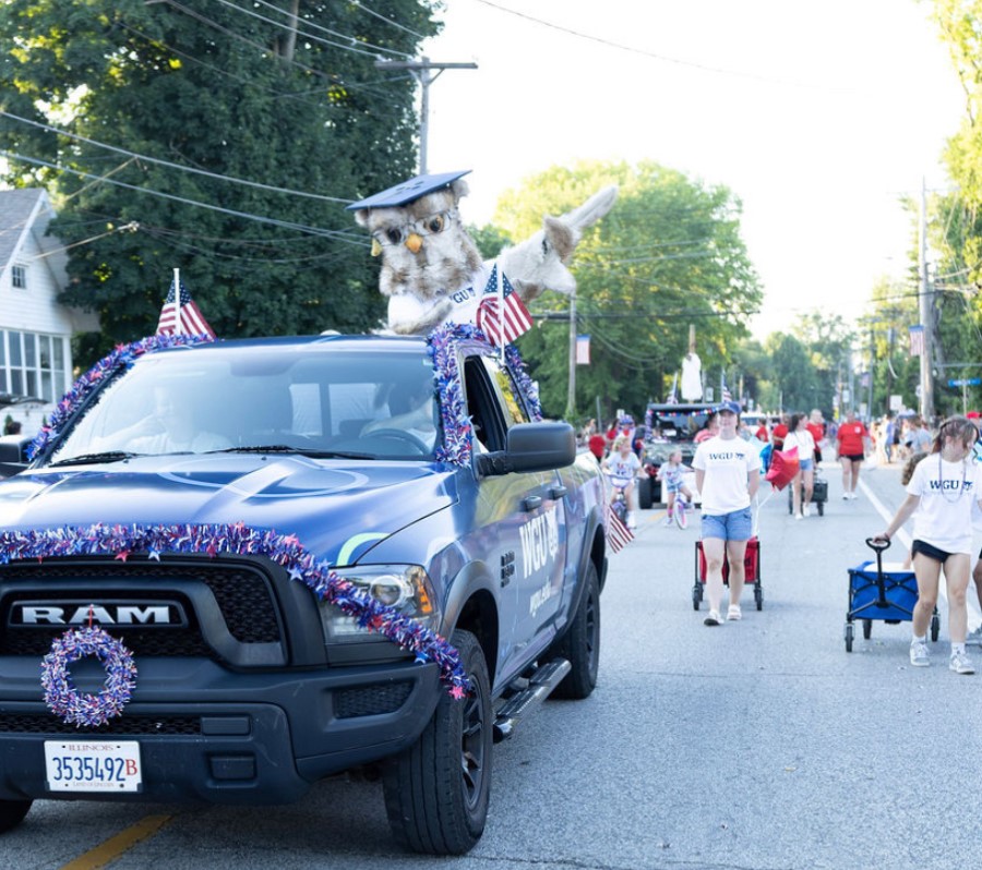 A blue pickup truck decorated with red, white, and blue garlands and American flags, featuring a large owl mascot wearing a graduation cap on its roof, participating in a parade.