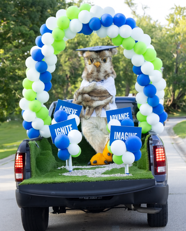 A mascot owl in a graduation cap and glasses stands in the back of a truck decorated with a green, blue, and white balloon arch, surrounded by signs reading "ACHIEVE," "ADVANCE," "IGNITE," and "IMAGINE."