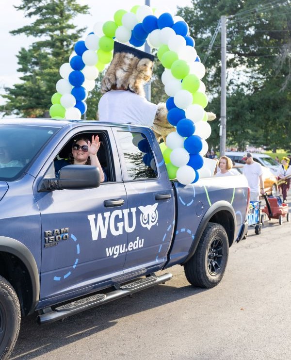 A WGU branded pickup truck with a mascot and balloons in the back, a woman is waving from the driver's seat.