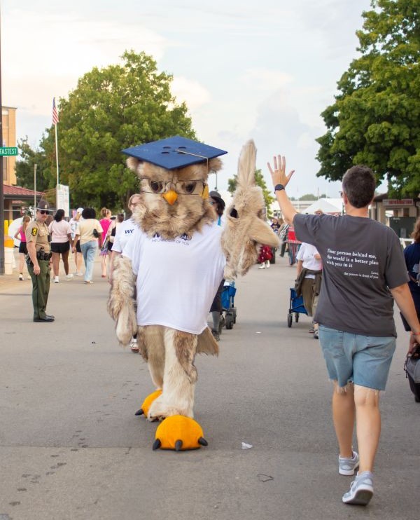 An owl mascot in a graduation cap and glasses walks down a street, wearing a white t-shirt that says "THE UNIVERSITY OF YOU".