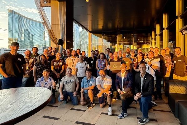 A large, diverse group of people posing for a photo on an outdoor patio with a city skyline in the background, one person holding a framed award.