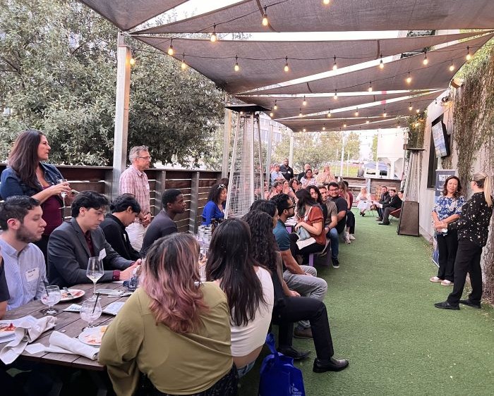 A large group of people gathered at an outdoor patio event with two women presenting to the crowd.