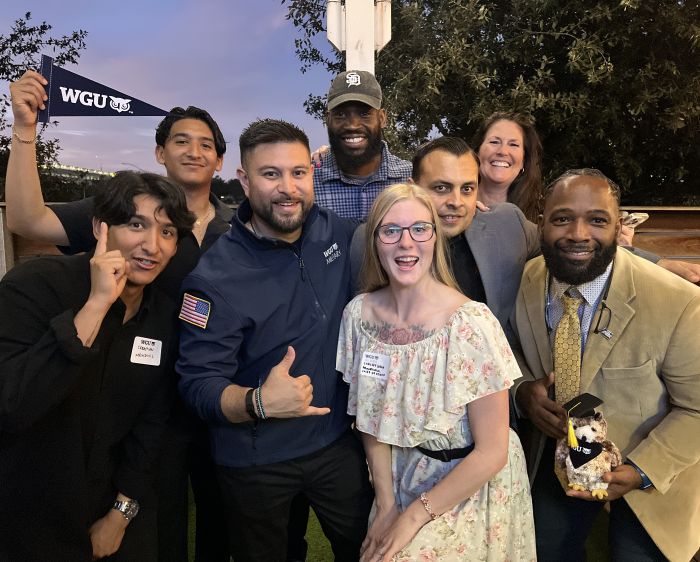 A diverse group of eight smiling individuals, some holding WGU branded items, pose together outdoors at an evening event.