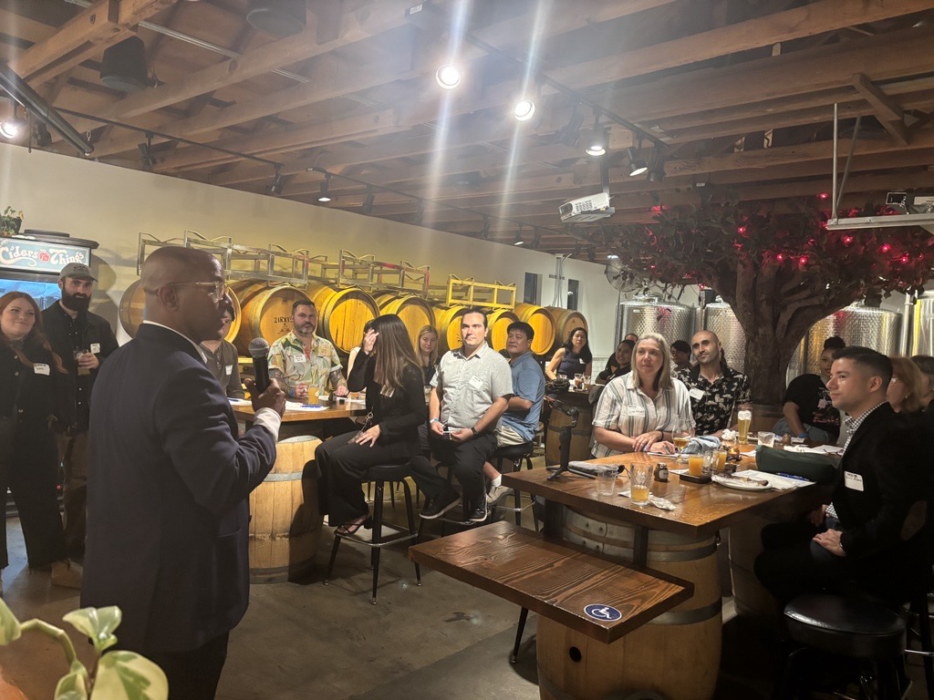 A speaker addresses an attentive audience in a brewery, with large wooden barrels lining the background.
