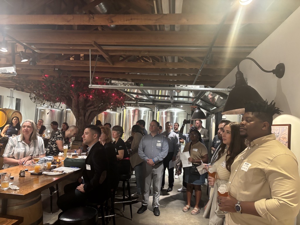 A diverse group of people gathered at an event in a brewery, with large stainless steel tanks in the background.