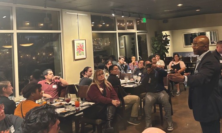 A man in a suit jacket speaks to a diverse group of people seated at tables in a dimly lit event space.