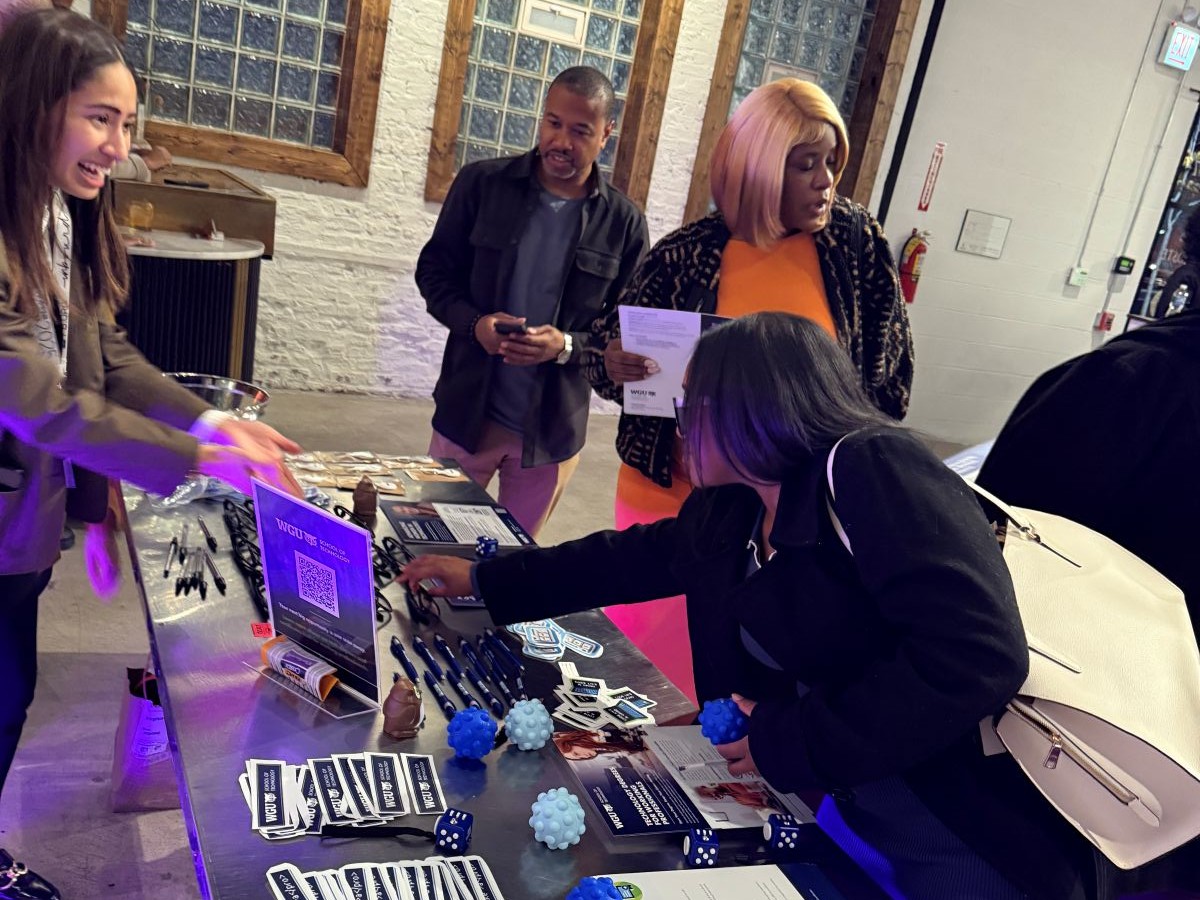 Attendees engaging at a WGU School of Technology event booth, with promotional materials and an attendant smiling.