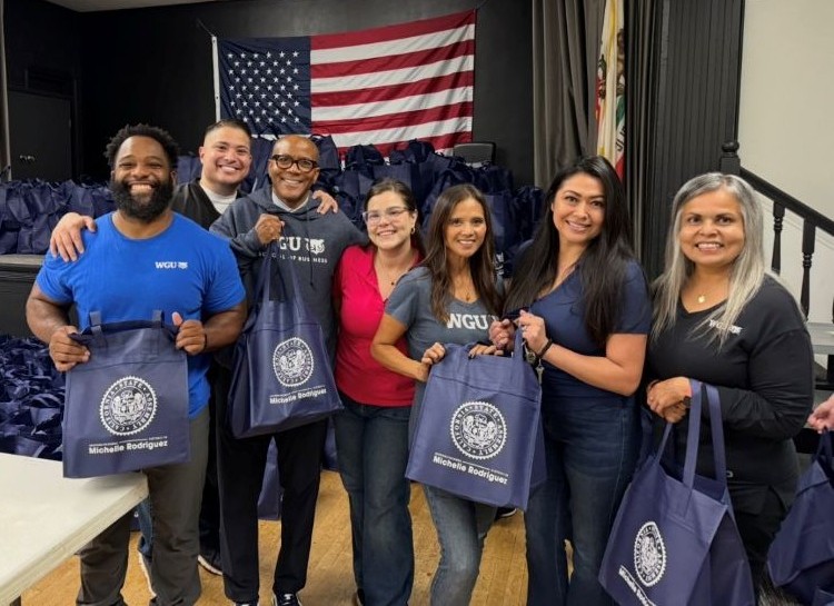 A diverse group of seven smiling people holding blue tote bags with a seal and the name "Michelle Rodriguez" printed on them, an American flag is in the background.