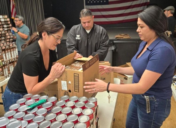 Three people organizing boxes and canned goods at a community event, with an American flag in the background.