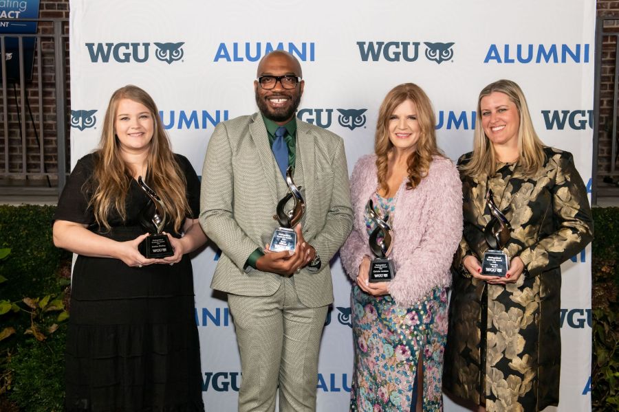 Four WGU alumni, three women and one man, smiling and holding awards at an event with a 'WGU ALUMNI' backdrop.
