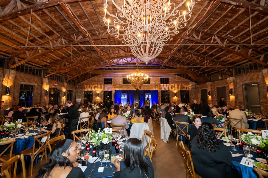 A formal dinner event in a large venue with a high wooden ceiling, chandeliers, and brick walls. Guests are seated at tables with blue tablecloths, with a stage displaying 'WGU' logos in the background.