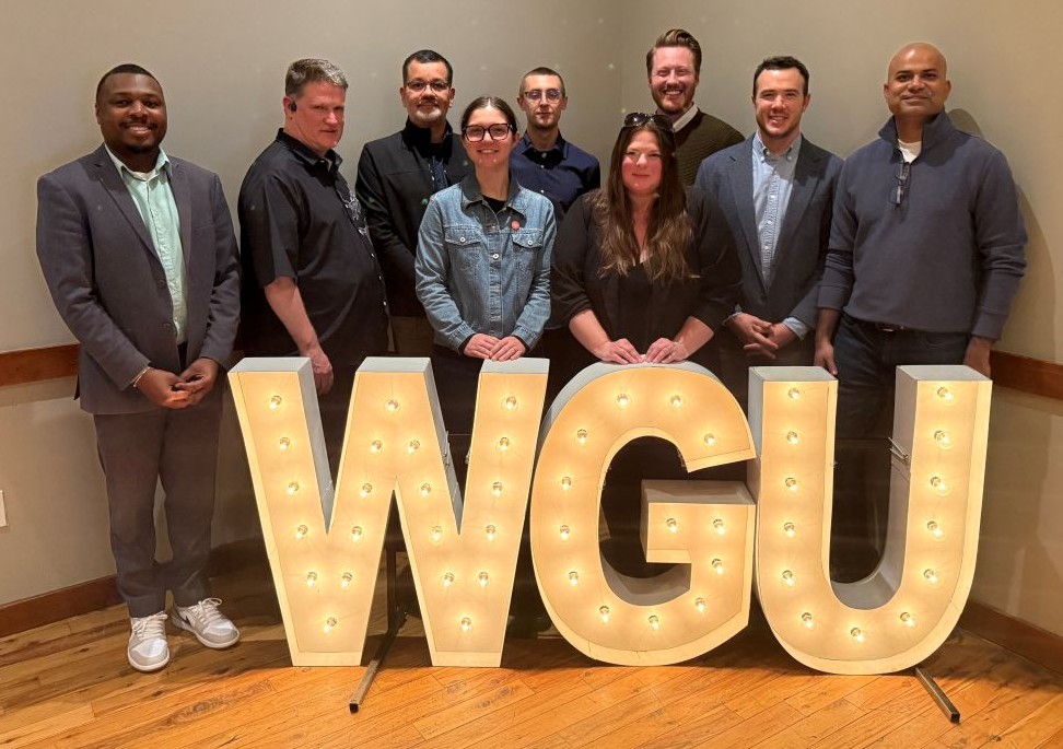 A diverse group of individuals, including seven men and two women, stand behind large, illuminated letters spelling "WGU".