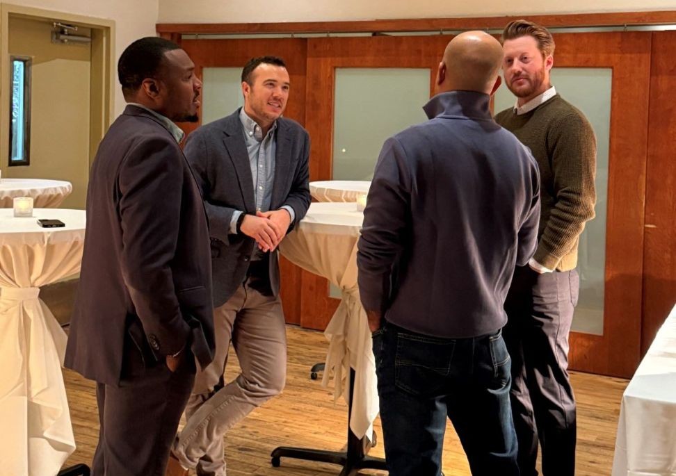 Four men conversing in a well-lit room with high-top tables, suggesting a business or social event.