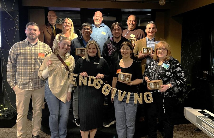A group of diverse people smiling and holding a glittery "Friendsgiving" banner, with several individuals also holding small gift boxes.