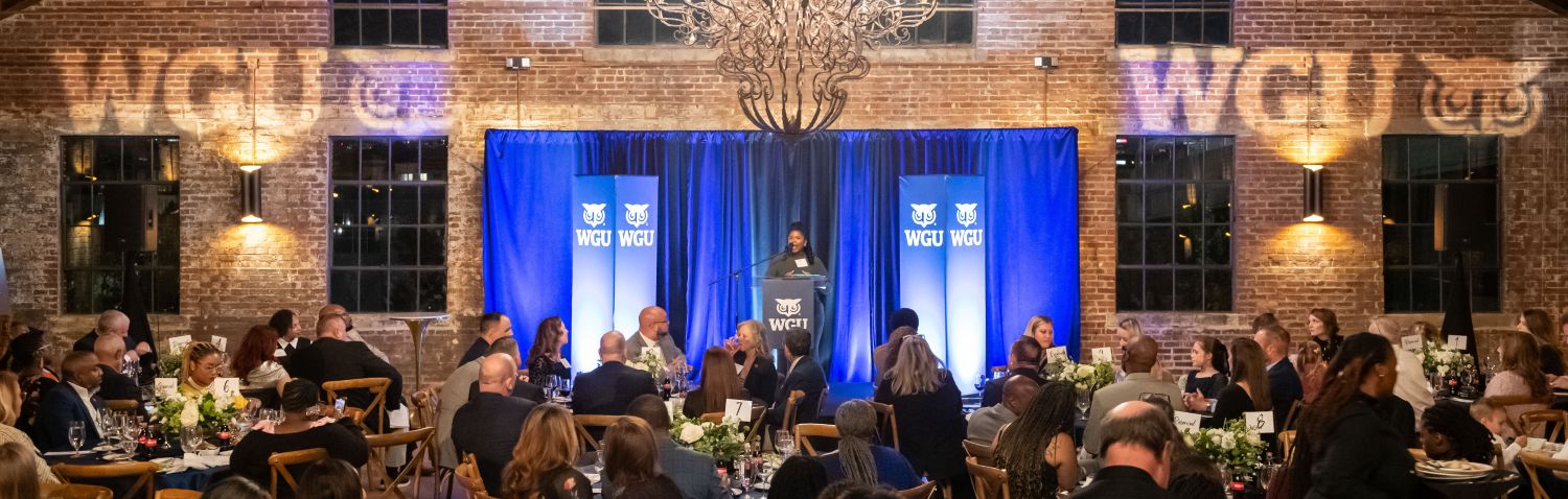 A person speaking at a podium on a stage with a blue WGU backdrop, facing an audience seated at elegant round tables in a brick-walled event hall.