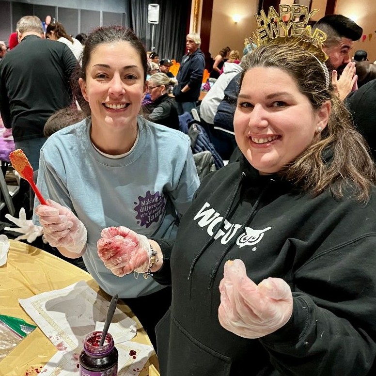 Two smiling women in gloves, one with a 'Happy New Year' headband, making a sandwich at an event.