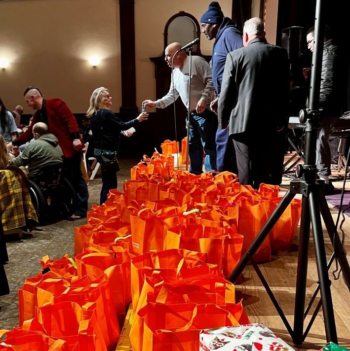 A large room filled with people and hundreds of orange reusable bags on a stage, with Christmas presents in the foreground.