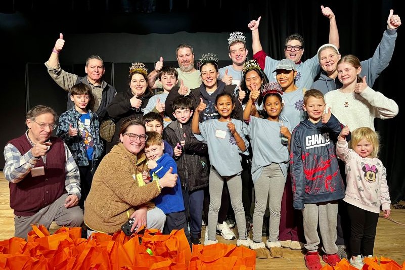 A large group of smiling adults and children on a stage, giving thumbs up, with many orange bags in the foreground.