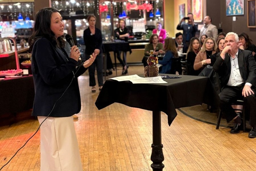 A speaker addresses a group of people at an indoor event, gesturing with her hand as she talks.