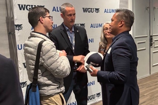 Attendees engaging in conversation in front of a branded WGU Alumni backdrop, with one person holding a university cap.