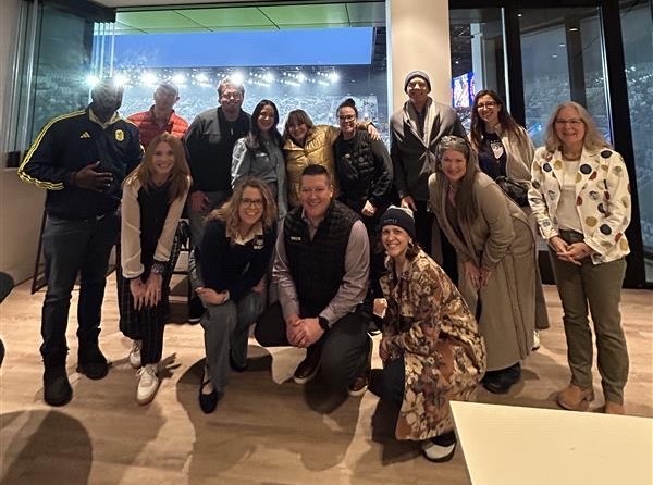 A diverse group of smiling people pose for a photo in an indoor suite overlooking a brightly lit stadium at night.