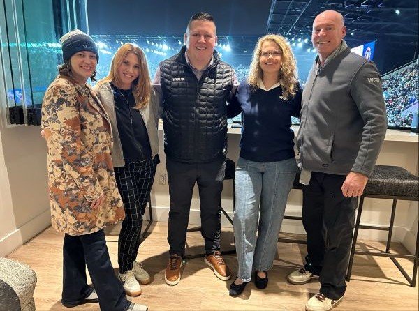 Happy group of five, including individuals in WGU gear, standing in an indoor setting with a large window revealing a lit stadium.