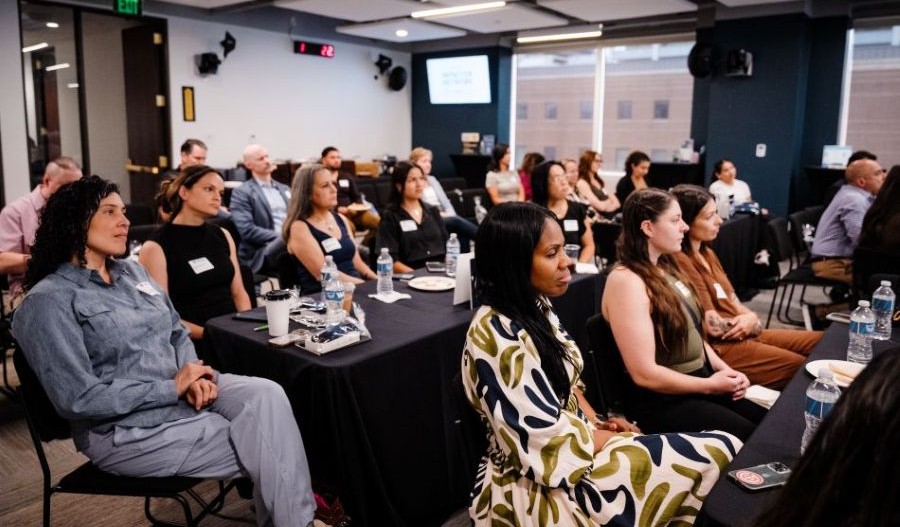 A diverse group of attendees, mostly women, seated at tables in a conference room, looking towards the front.