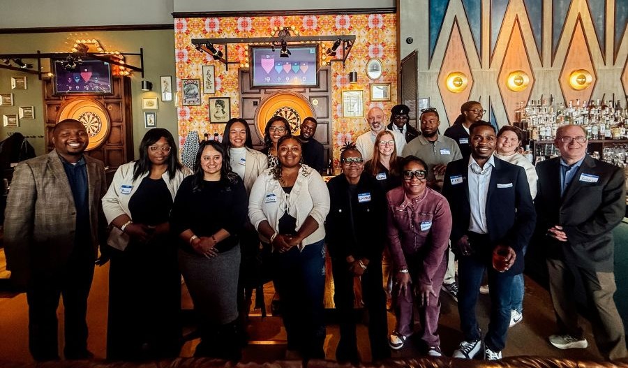A diverse group of smiling professionals and attendees gathered in a social venue, with a dartboard and bar in the background.
