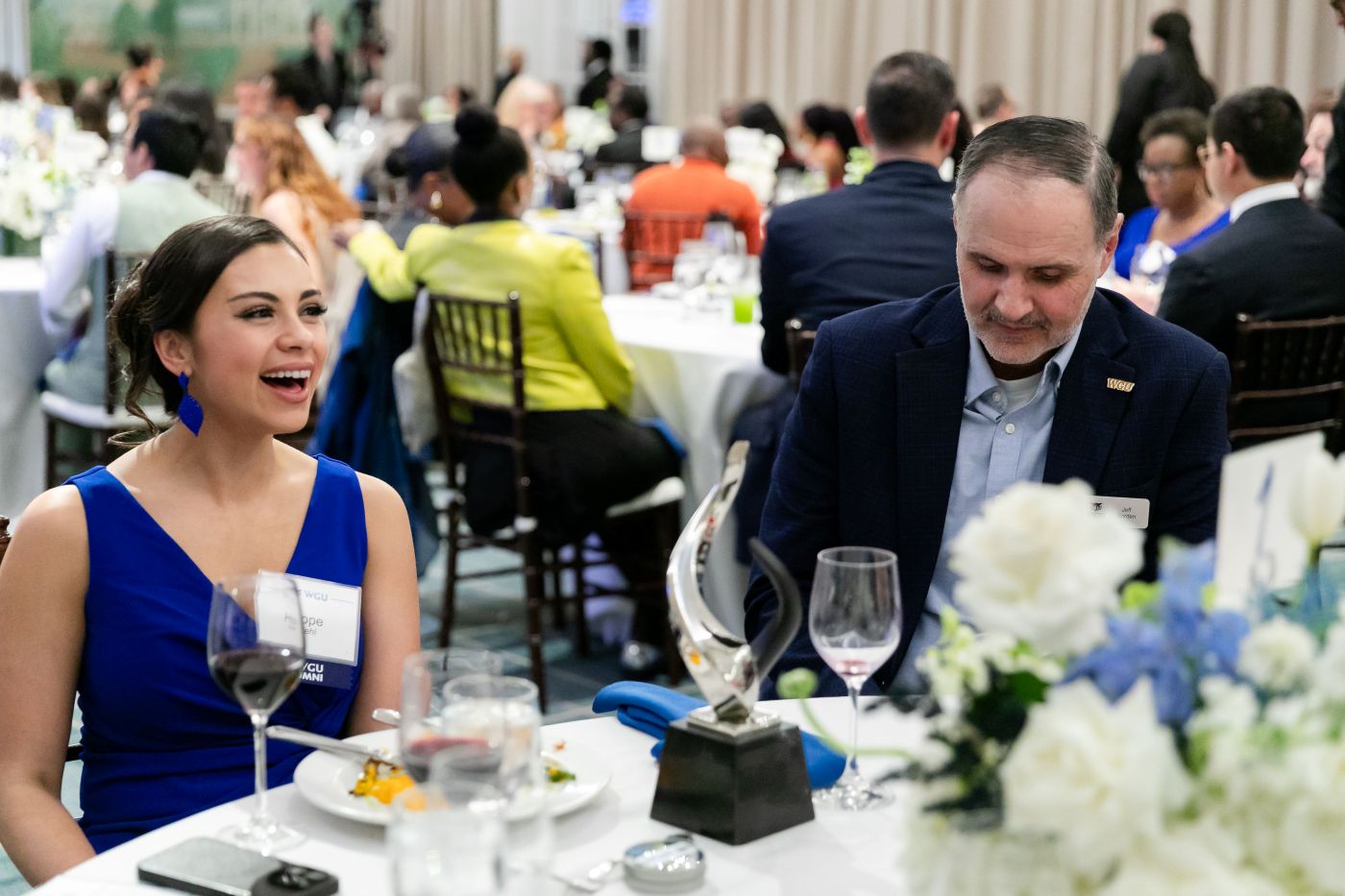 A woman in a blue dress laughs while sitting at a formal dinner table next to a man in a suit, with a silver award and floral centerpiece between them.