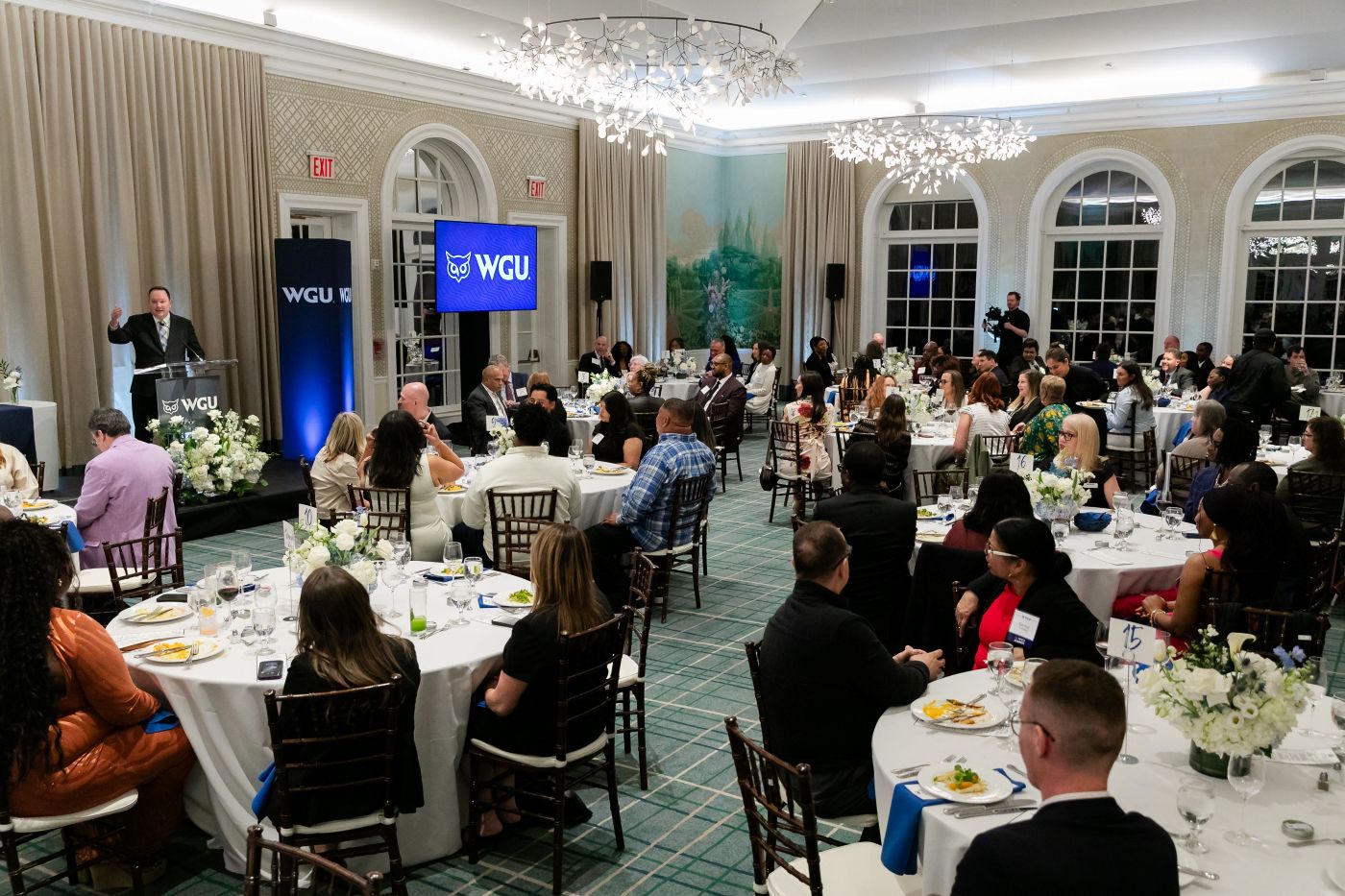 A speaker at a podium addresses a large audience seated at round tables in an elegant ballroom, with a screen displaying the WGU logo.