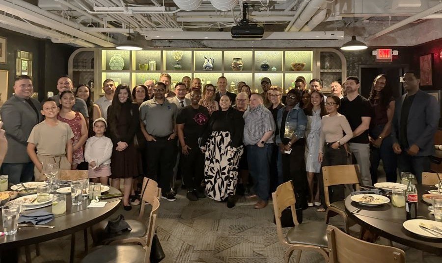 A diverse group of adults and children smiling and posing together in a restaurant setting with tables and decorative shelves in the background.