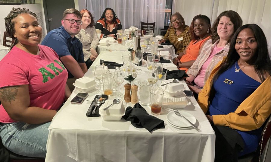 A diverse group of smiling people gathered around a long table in a restaurant, with drinks and takeout containers.