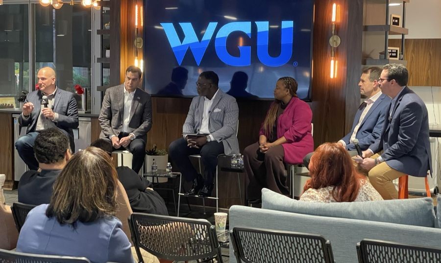 Panel discussion with six speakers in front of a blue WGU logo, one man speaking into a microphone, and an audience listening intently.