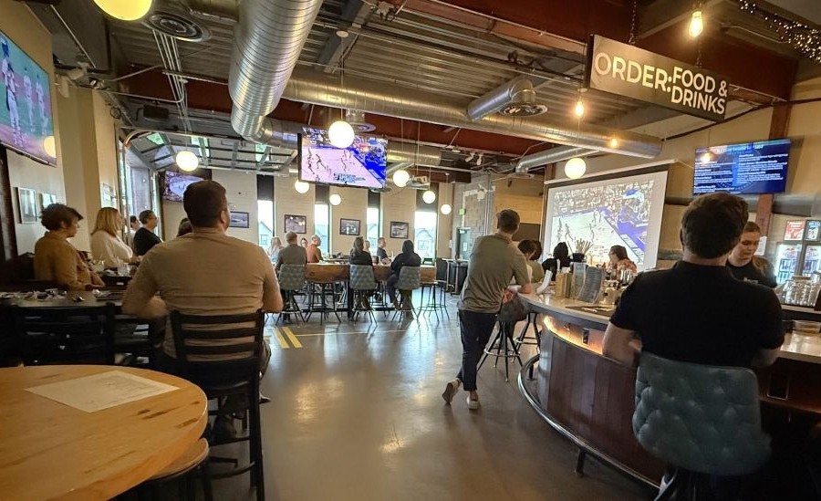 Interior view of a lively bar or restaurant with patrons seated at tables and a bar, watching sports on multiple screens and a large projector.