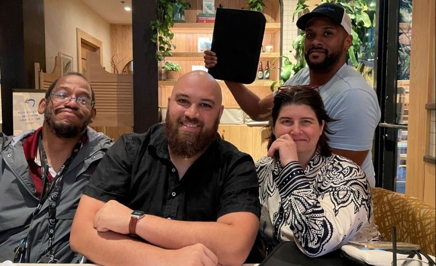 Four smiling people at a table, with one man holding up a black folder in the background.