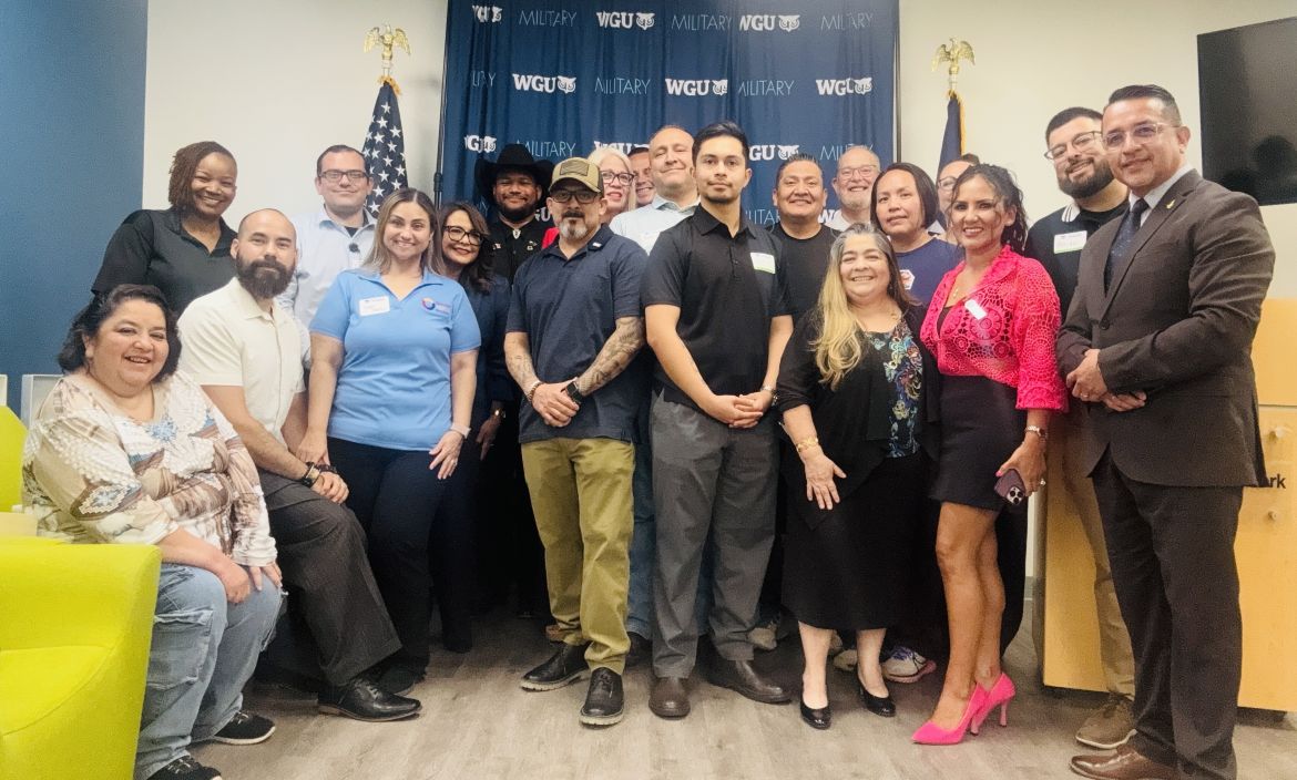 A diverse group of smiling people, including men and women, pose together in front of a banner that reads 'WGU MILITARY'.