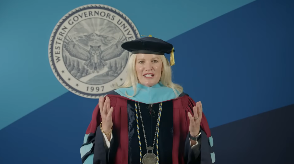 A blonde woman in academic regalia, including a cap, gown, and hood, speaking in front of the Western Governors University seal.