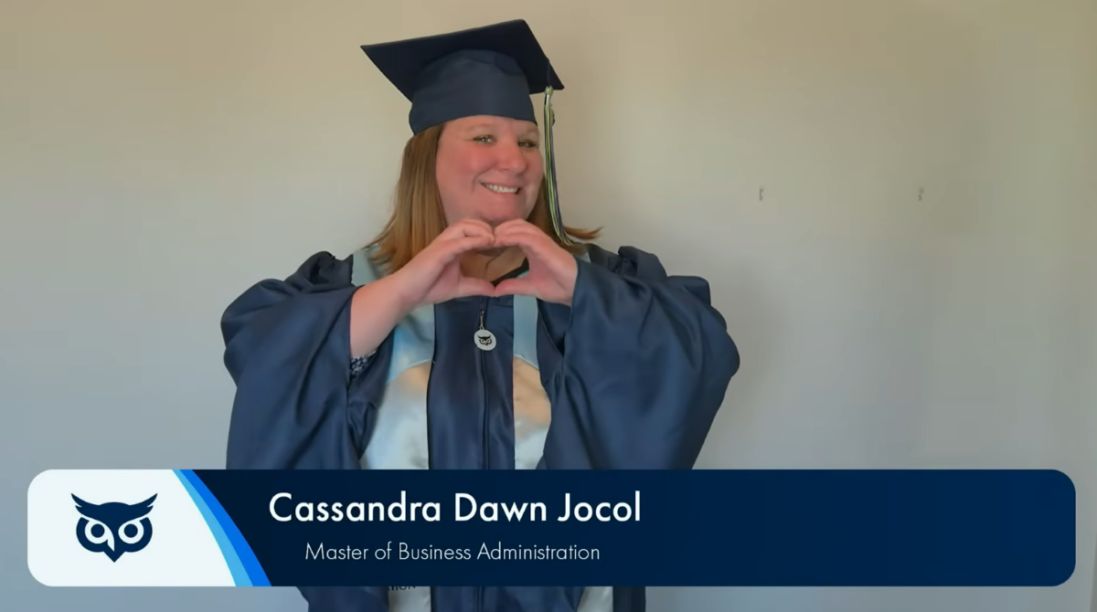 Cassandra Dawn Jocol, a graduate in a cap and gown, smiles while making a heart shape with her hands.
