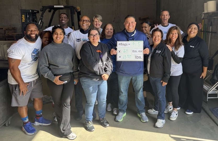 A diverse group of smiling people stands together in a warehouse, one person holding a whiteboard
