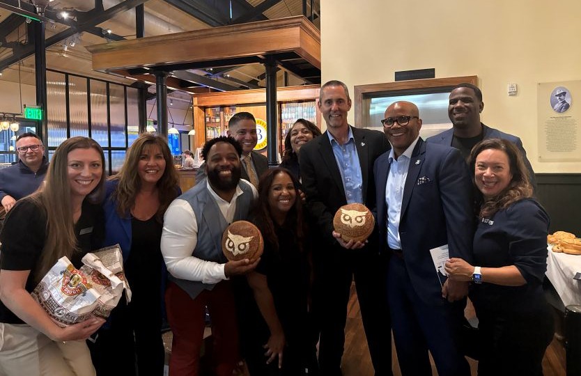 A diverse group of smiling professionals poses indoors, with several holding round, bread-like items featuring an owl logo.