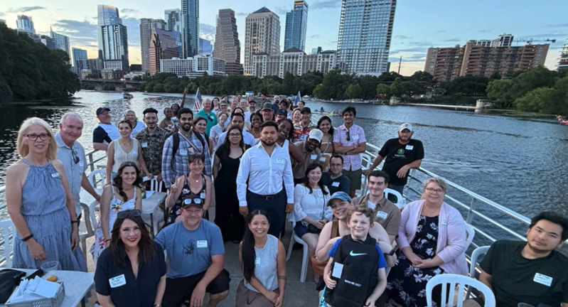 A large, diverse group of people smiling on a boat, with a city skyline visible in the background.
