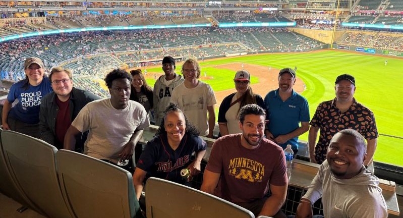 A diverse group of people smiling and posing for a photo at a baseball stadium, with the game in progress on the field below.