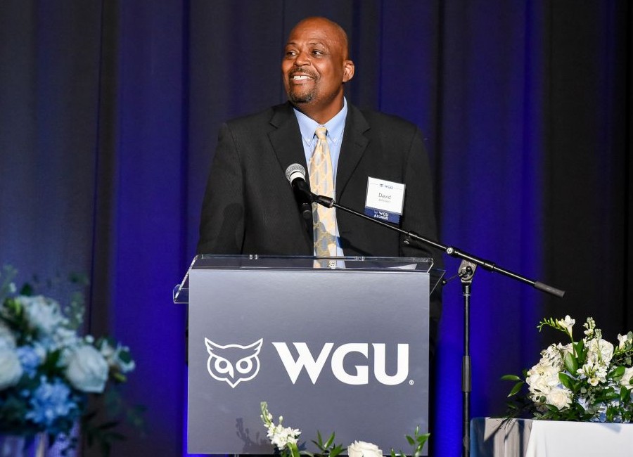 A smiling man, identified as David, speaks at a podium with the WGU logo, dressed in a suit and tie, against a blue curtain backdrop.
