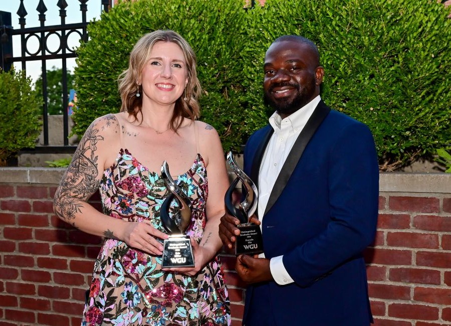 A smiling woman and man, both holding silver and black awards, stand outdoors in front of green bushes.