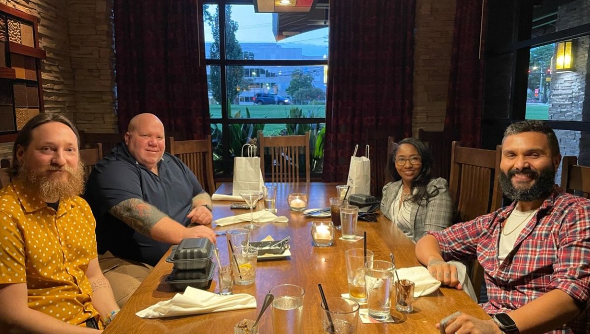 Four colleagues smile around a candlelit restaurant table set with drinks, plates, and small gift bags.
