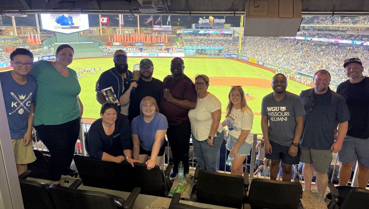 A diverse group enjoys a baseball game from a covered suite, standing and seated above the field.